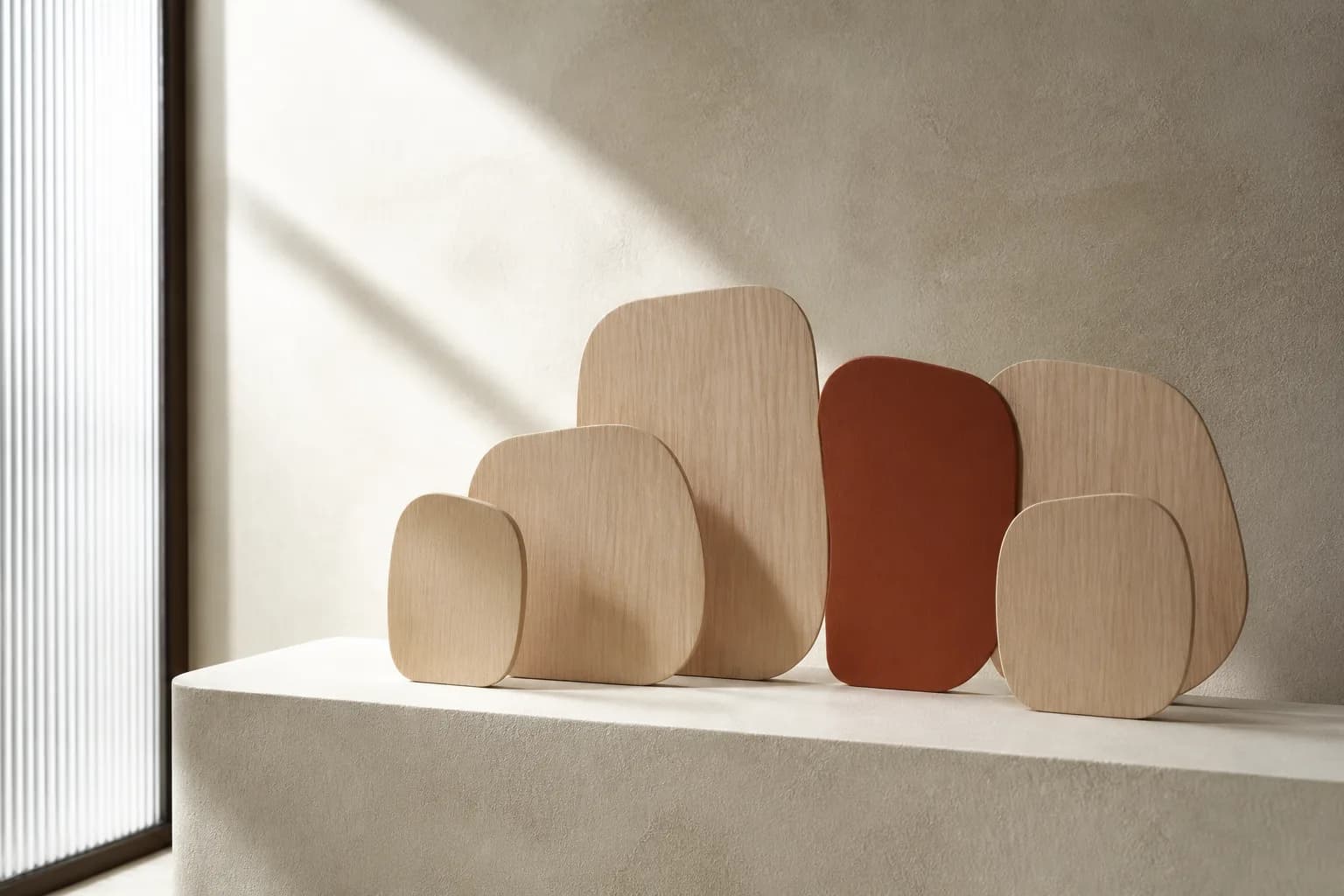 Nested pale oak plates arranged like overlapping speech forms on a cream plinth, one in terracotta.